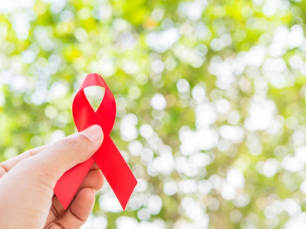 Concepto del Día Mundial del SIDA. Mano sosteniendo cinta roja sobre hojas de árbol fondo bokeh . — Foto de Stock