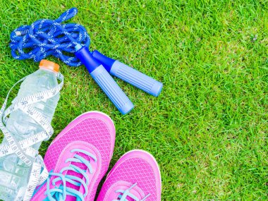 Çift pembe spor ayakkabı, havlu, kulaklık, su şişesi ve yeşil çim sahada ip atlama. İçinde arka plan orman veya park iz. Spor çalıştırmak için aksesuarlar.