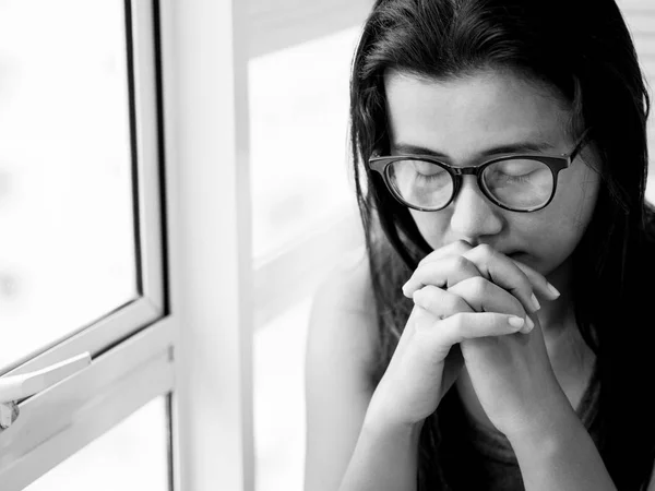 sad woman wear glasses sitting alone and praying near the window.