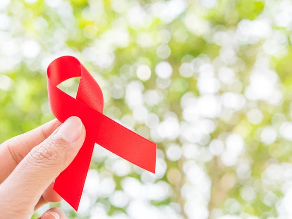 Concepto del Día Mundial del SIDA. Mano sosteniendo cinta roja sobre hojas de árbol fondo bokeh . — Foto de Stock