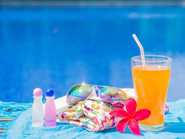 Frangipani rojo (plumeria) flores, hierbas solares, champú, loción, sombrero, toallas enrolladas y jugo de naranja con un trozo de limón al lado de la piscina. Vacaciones, playa, concepto de viaje de verano — Foto de Stock