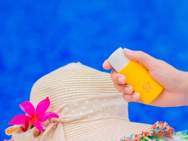 UV güneş kremi şişeyle yaz şapka ve kırmızı frangipani Yüzme Havuzu tarafından (plumeria) çiçek tutan kadın el. Tatil ve dinlenme, yaz seyahat kavramı.