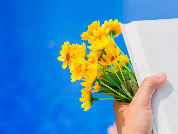 スイミング プールのそばの本と黄色い花を持つ女性の手。休暇とリラックス、夏旅行の概念. — ストック写真