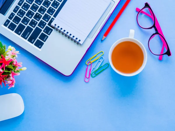 Feminino espaço de trabalho de mesa com acessórios de escritório, incluindo laptop, livro de notas, lápis vermelho, mouse, clipes, óculos rosa e flor no fundo azul . — Fotografia de Stock