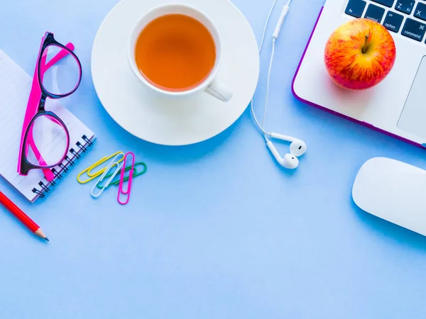 Top view of feminine desk workspace with office accessories including laptop, note book, red pencil, red apple, pink glasses and coffee on blue background. and coffee on blue background.