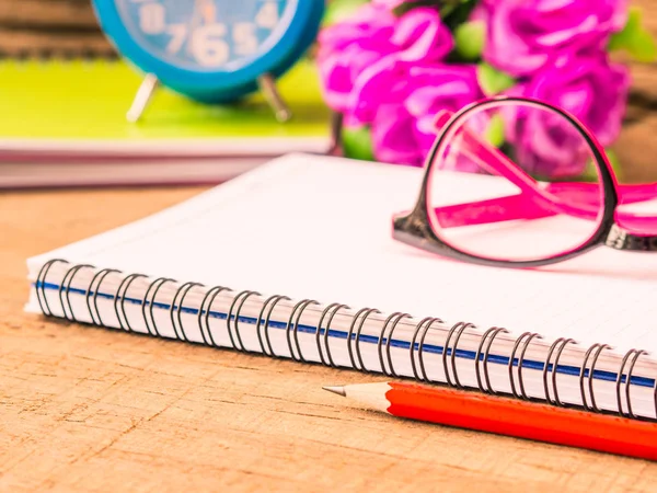 Toebehoren voor kantoor met inbegrip van de nota boek, rood potlood, roze bril en wekker op houten achtergrond. Onderwijs en bedrijfsleven concept. — Stockfoto