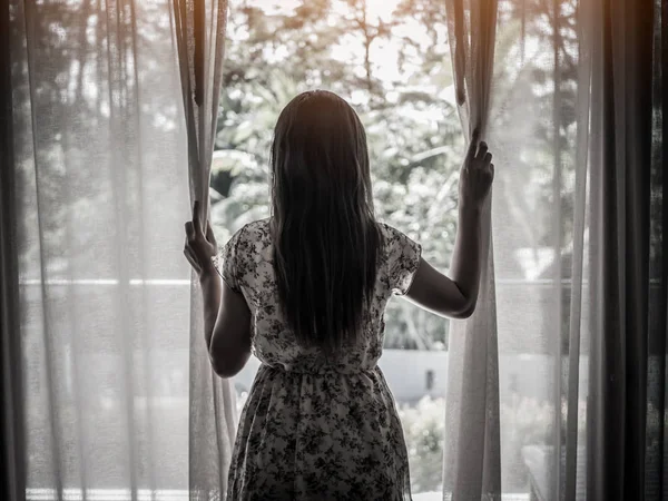 Bakifrån av en ung kvinna hålla gardinerna öppna att titta genom fönstret. Hopplös och ledsen kvinna koncept. — Stockfoto