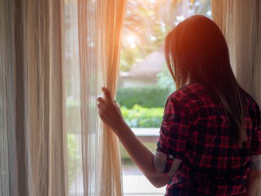 Evde bir büyük ışık pencereden dışarı bakmak için perdeleri açık tutan genç kadın.