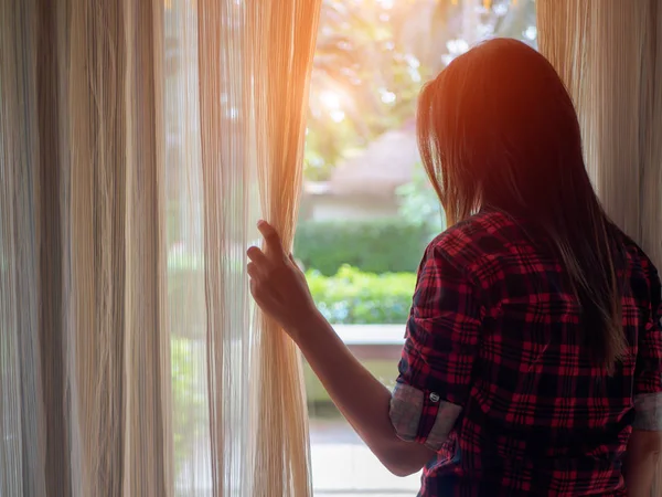 Evde bir büyük ışık pencereden dışarı bakmak için perdeleri açık tutan genç kadın.
