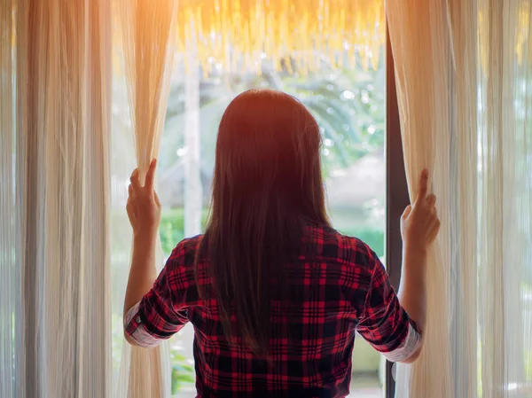 Evde bir büyük ışık pencereden dışarı bakmak için perdeleri açık tutan genç kadın.