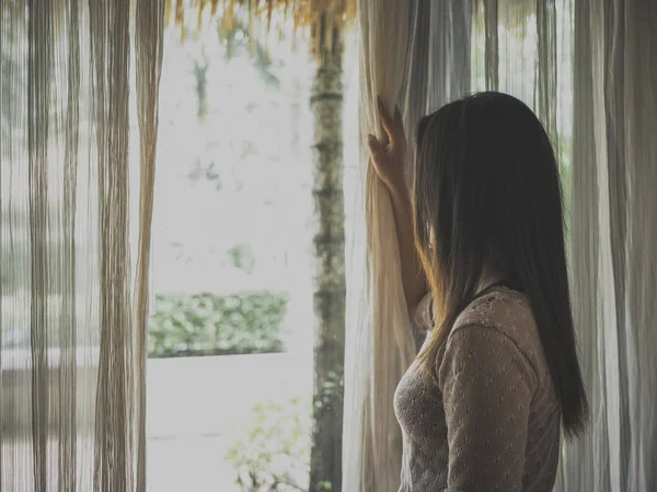 Sad woman holding the curtains open to look out of a large light window at home.