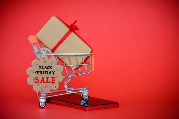 Online-Shopping-Konzept. Warenkorb, Handy- und Geschenkbox mit schwarzer Freitagsverkaufskarte auf rotem Hintergrund. — Stockfoto