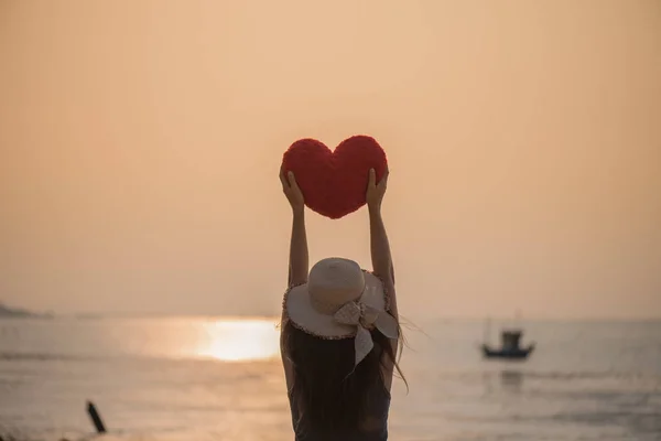 Женщина держит в руках красную подушку в форме сердца на пляже во время заката на День Святого Валентина и концепция любви . — стоковое фото