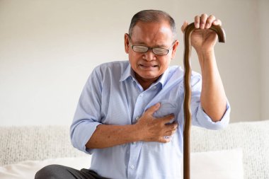 Asian old man sitting on sofa and having a with pain on heart, heart attack at home. Senior healthcare concept. clipart