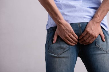 Genç adam karın ağrısı ve hemoroit olduğu için poposunu tutuyor. Sağlık hizmeti konsepti. Basur ve kabızlık çeken bir adam..