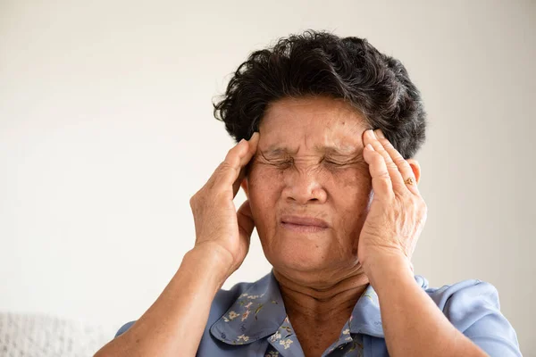 Asian old woman sitting on sofa and having a headache at home. Senior healthcare concept.