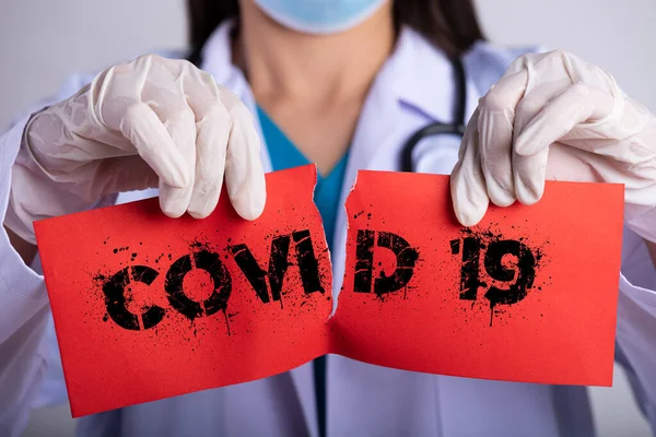 Female doctor wearing surgical mask, stethoscope and holding red torn paper with COVID-19 text. Healthcare and medical concept.
