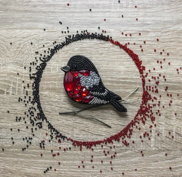 A handmade brooch made of beads and felt in the form of a bullfinch bird lies on a wooden background surrounded by beads of red and black color