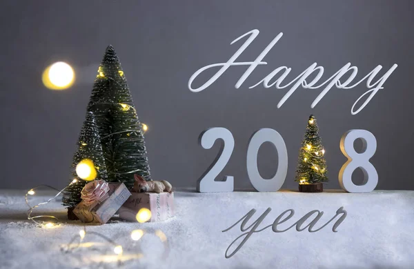Sur un terrain enneigé sous les sapins, le chien dort sur des cadeaux et au loin sont les chiffres 2018 où dans le rôle d'un arbre de Noël avec carte de lumières heureux 2018 année — Photo
