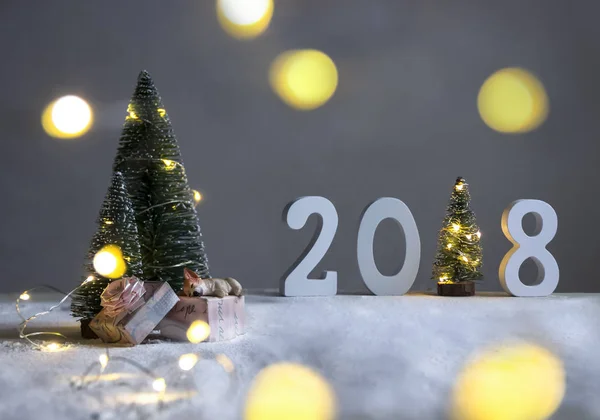 Sur un terrain enneigé sous les sapins, le chien dort sur des cadeaux et au loin sont les chiffres 2018 où dans le rôle d'un arbre de Noël avec des lumières — Photo