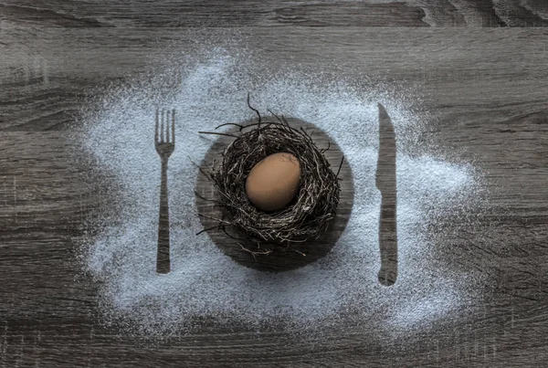Sobre un fondo de madera con nieve en polvo en polvo cubierto con una silueta un plato un cuchillo de tenedor de electrodomésticos en el centro se encuentra un huevo en un nido — Foto de Stock