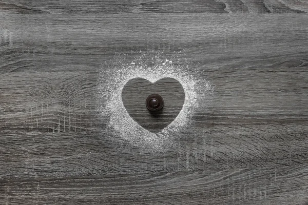 På en trä bakgrund med pulveriserad pulver, en siluett av hjärtat hälls med snö och i mitten ligger choklad runda godis — Stockfoto