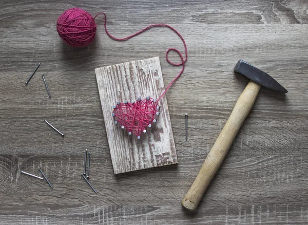 Op een houten tafel ligt een hamer van een nagel van een roze draad en een cadeau met je handen een hart van draden en nagels — Stockfoto