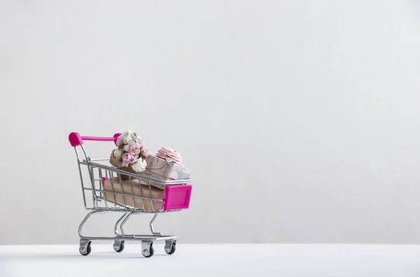 No chão branco em um fundo branco há um carrinho com um presente rosa com um arco e um buquê de flores rosas — Fotografia de Stock