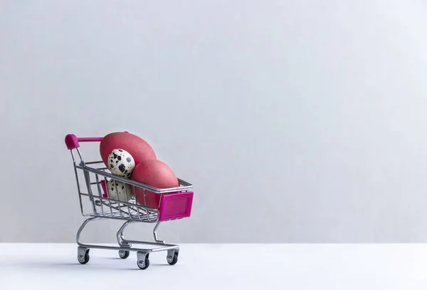 No chão branco em um fundo branco há um carrinho de compras com ovos de codorna e frango vermelho — Fotografia de Stock