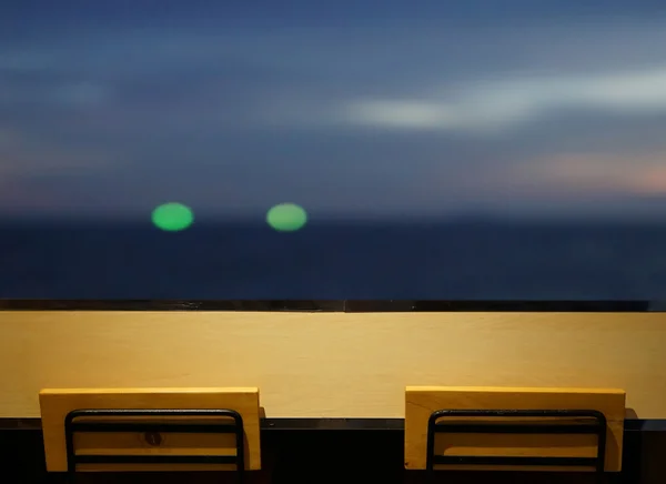 Holz lange Tischplatte mit 2 einfachen Stühlen am Fenster mit Meerblick in der Nacht mit Bokeh-Licht von Fischerboot weit weg als Hintergrund — Stockfoto