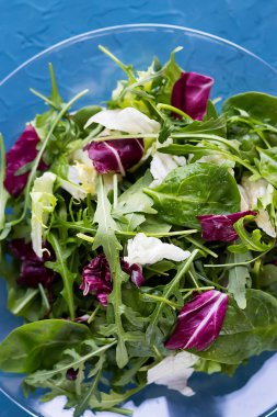 Taze roka salatası, ıspanak, buzdağı marulu, zencefilli ve limonlu salata mavi arka planda şeffaf bir tabakta. Yumuşak odaklanma. Vejetaryenlik ve çiğ gıda diyeti kavramı.