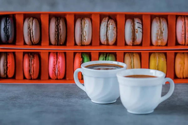 Assorted macaroon cakes in a red box and two white cups of coffee on a gray background. Soft focus.