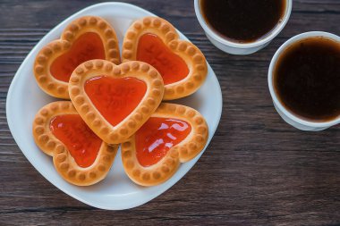 Beyaz tabakta çilek reçelli kalp şeklinde kurabiyeler ve kahverengi ahşap arka planda iki beyaz kahve. Aşk konsepti. Yukarıdan bak. Yumuşak odak.