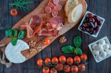 Prosciutto, ekmek, zeytin, ceviz, mozzarella, salam, fesleğen ve kiraz domatesleri kahverengi ahşap tahtada. Akdeniz mutfağı. Üst görünüm