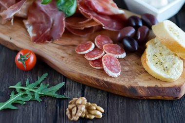Prosciutto, ekmek, zeytin, ceviz, mozzarella, salam, fesleğen ve kiraz domatesleri kahverengi ahşap tahtada. Akdeniz mutfağı. Yumuşak odak.