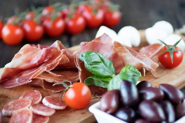 Prosciutto, ekmek, zeytin, ceviz, mozzarella, salam, fesleğen ve kiraz domatesleri kahverengi ahşap tahtada. Akdeniz mutfağı. Yumuşak odak.