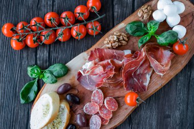 Prosciutto, ekmek, zeytin, ceviz, mozzarella, salam, fesleğen ve kiraz domatesleri kahverengi ahşap tahtada. Akdeniz mutfağı. Yumuşak odak.