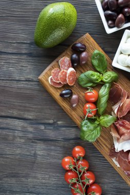 Prosciutto, zeytin, salam, fesleğen, avokado ve vişneli domatesler kahverengi ahşap tahtada. Akdeniz mutfağı. Yumuşak odak.