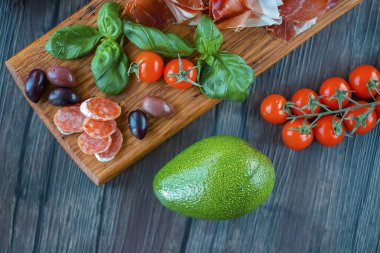 Prosciutto, zeytin, salam, fesleğen, avokado ve vişneli domatesler kahverengi ahşap tahtada. Akdeniz mutfağı. Yumuşak odak.