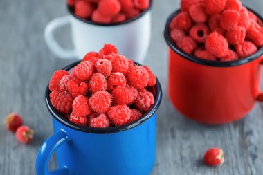 Gri ahşap arka planda mavi, kırmızı ve beyaz metal bardaklarda organik ahududular. Yumuşak odak.