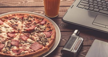 Pizza üzerine bir tabak, siyah dizüstü bilgisayar, elektronik sigara veya vape, cep telefonu ve bir bardak meyve suyu ahşap tablo