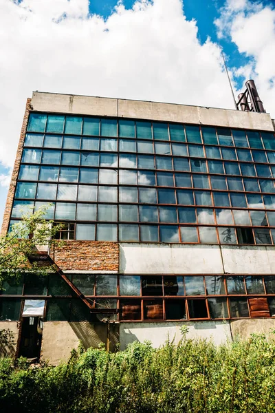 Large abandoned factory exterior, overgrown with plants, large windows with reflection of clouds and sky