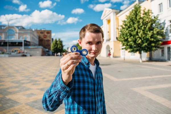 Молодой человек в голубой рубашке показывает Фиджета Спиннера, избирательный фокус на Спиннере с размытым фоном — стоковое фото