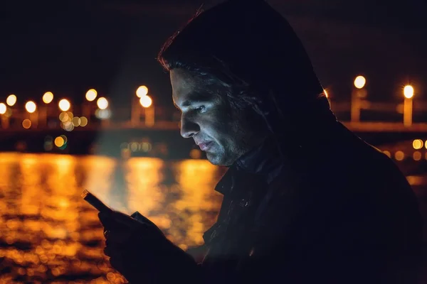 Homem com smartphone à noite no fundo embaçado das luzes da cidade refletidas na água — Fotografia de Stock