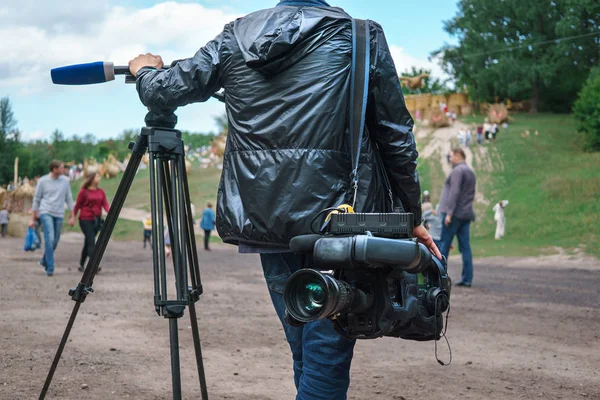 Professional video operator or videographer standing with tripod and digital camera at blurred background of park with walking people — Stock Photo, Image