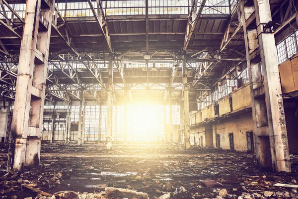 Inside abandoned large industrial warehouse hall with garbage and columns, sunset light shining through big windows — Stock Photo, Image