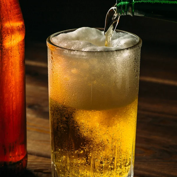 Pouring foaming beer into glass mug with drops near cold beer bootle, close up