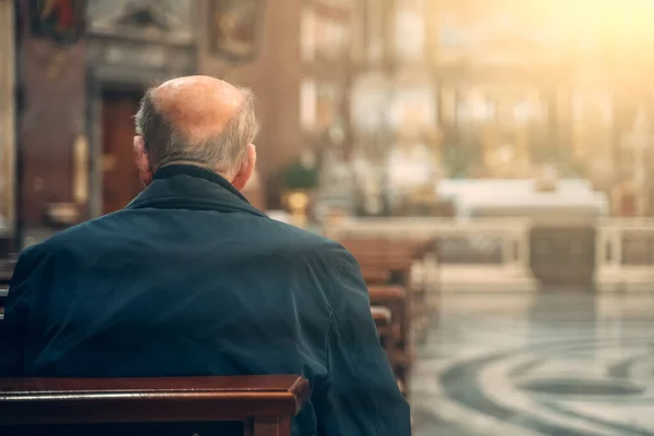 Elderly man prays while sitting on bench inside church, rear view — Stock Photo, Image