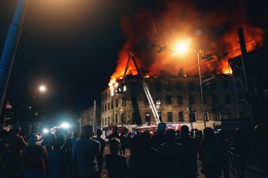 Gece yanan şehir binası, itfaiyeciler evi, sivillerin siluetlerini ön plandaki insanları ve yanan ateşi dumanla söndürüyor.