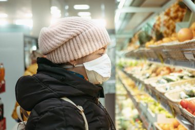 Süpermarketteki tıbbi maskeli genç kadın meyve ve diğer yiyecekleri seçiyor. Coronavirüs salgını Covid-19 virüsü